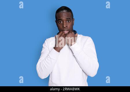A young handsome man wearing white sweater over blue background covers mouth and looks with wonder at camera, cannot believe unexpected rumors. Stock Photo