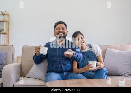 Indian couple watch tv on the couch at their living room, husband and wife spending quality time together pleasure smile. man holding remote control Stock Photo