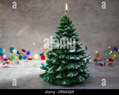 A lit Christmas tree shaped candle against the bokeh of a colourful fairy light decoration with a concrete grey background and space for copy Stock Photo