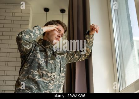 Serious short-haired young man in military uniform soldier looking through the window, upset soldier suffering from posttraumatic syndrome after Stock Photo
