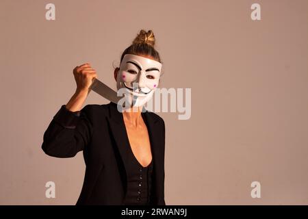 Woman with an anonymous mask dressed in a black colored suit jacket, carrying a knife in her hand, prepared for the Halloween party Stock Photo