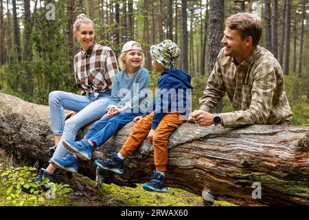 happy family with kids sitting and resting on fallen tree after walk in forest. outdoor activities, nature adventure Stock Photo