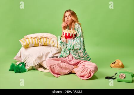 Full length portrait of terrified woman in pajama watchingTV programme, horror film holding pop corn against on pastel green background. Stock Photo
