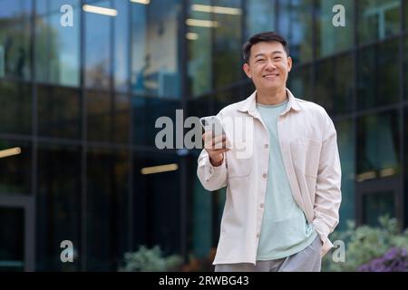 Young Asian man businessman, freelancer, designer standing outside office buildings, using mobile phone and smiling at camera. Stock Photo