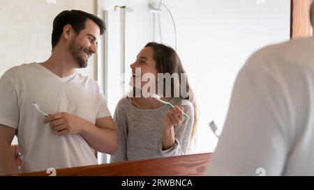 Laughing couple cleaning teeth, holding toothbrushes, standing near mirror Stock Photo