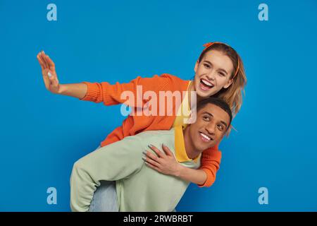 joyful african american man piggybacking cheerful girlfriend on blue background, couple having fun Stock Photo