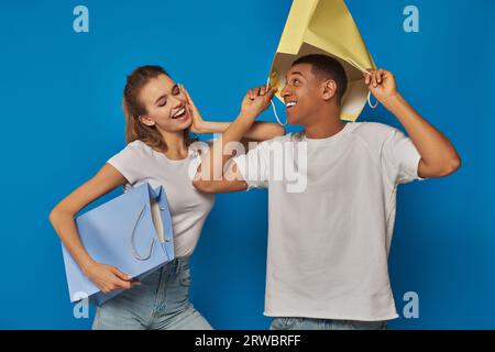 cheerful multicultural couple in casual attire holding shopping bags on blue backdrop, buying spree Stock Photo