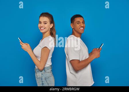 cheerful multicultural couple standing back to back and using smartphones on blue background Stock Photo
