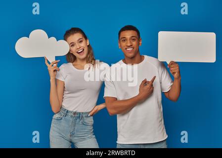 excited interracial couple pointing at blank placards on blue backdrop, thought and speech bubbles Stock Photo