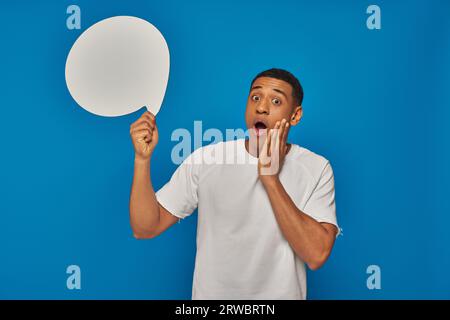 stunned african american man with open mouth holding blank speech bubble on blue background Stock Photo