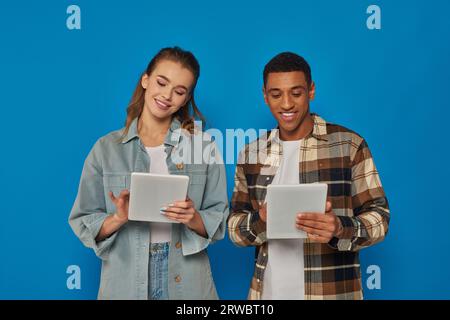 happy interracial man and woman using digital tablets on blue backdrop, social media and networking Stock Photo