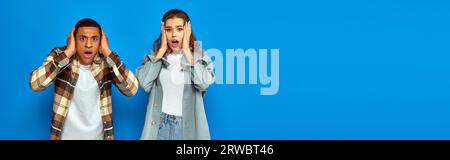 shocked interracial couple looking at camera on blue background, emotional reaction, banner Stock Photo