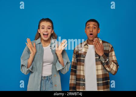 happy interracial couple screaming from joy looking at camera on blue backdrop, reacting emotionally Stock Photo