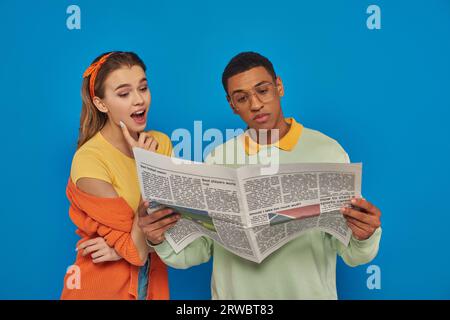 amazed woman reading newspaper near calm african american man in eyeglasses on blue backdrop Stock Photo