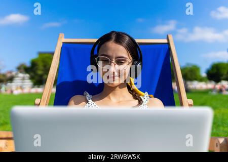 Happy young female in eyeglasses with laptop and headphones relaxing on chair in summer day and listening to music Stock Photo