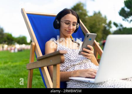Young positive female with eyeglasses in deckchair with laptop using mobile phone in park in summer day Stock Photo