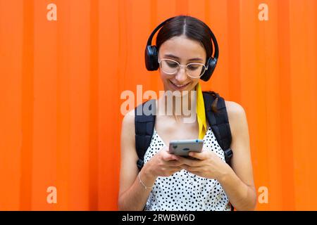 Happy young female in eyeglasses listening to music via headphones and browsing mobile phone against orange background Stock Photo