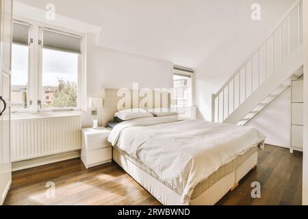 Interior of spacious bedroom with comfortable bed next to bright windows and staircase in modern apartment with white walls Stock Photo