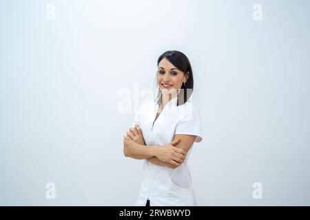 Happy young female doctor in medical uniform smiling and looking at camera while standing with folded arms against white background in hospital Stock Photo