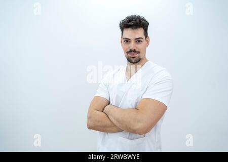 Serious young bearded male therapist in medical uniform crossing arms and looking at camera while standing against white background in light clinic Stock Photo