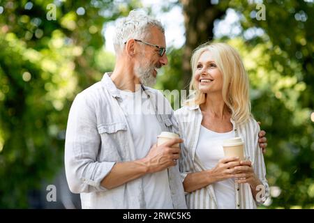 Retirement Leisure. Smiling Senior Spouses Walking Outdoors And Drinking Takeaway Coffee Stock Photo