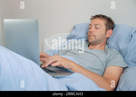 Male freelancer working with laptop in bed in the morning. Man designer on remote work from home. Wireless technology  Stock Photo