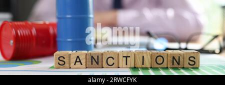 Wooden cubes with word sanction, oil drums and documents on table and msnager in background. Stock Photo