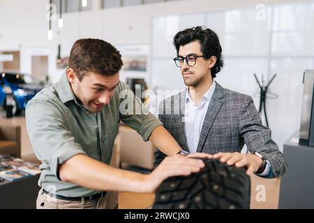 Confident salesman in business suit talking about characteristic of tires product to cheerful customer male came to look at assortment represented in Stock Photo