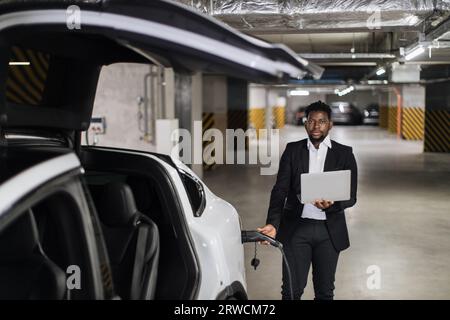 Man with computer plugging in EV charging cord in parking Stock Photo
