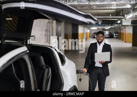 Man with computer plugging in EV charging cord in parking Stock Photo