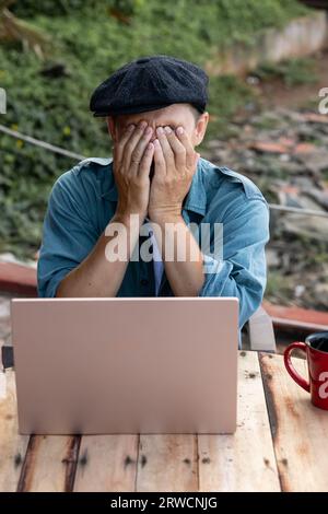 The man covered his face with his hands. A man in a hat is working on a laptop Stock Photo