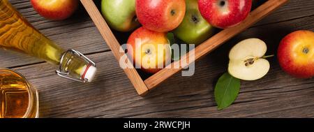 Ripe green and red apples in wooden box with branch of white flowers, glass and bottle of cider on a wooden table. Top view panoramic. Stock Photo