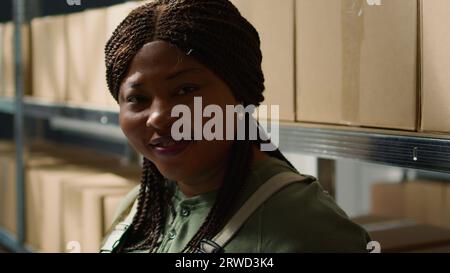 Portrait of smiling warehouse head of logistics holding digital tablet, checking packaging, ensuring flawless operations while employee in background handles packaged merchandise in cardboard boxes Stock Photo