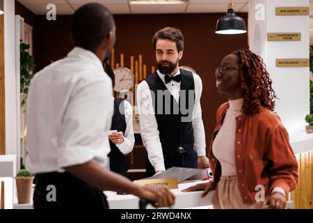 Hotel staff welcoming guests at counter, providing excellent check in service at hotel. Young employee talking to couple with luggage, looking for accommodation upon their arrival. Stock Photo
