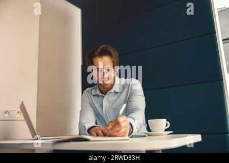 Smiling male freelancer making notes during working on laptop sitiing desk in coworking Stock Photo