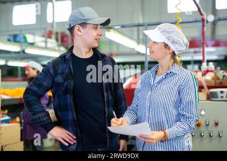 Workers of fruit storage and sorting facility talking about work process standing near the conveyor line and tangerine boxes. Stock Photo