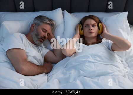 Irritated woman with headphones lying near her snoring husband in bed at home Stock Photo