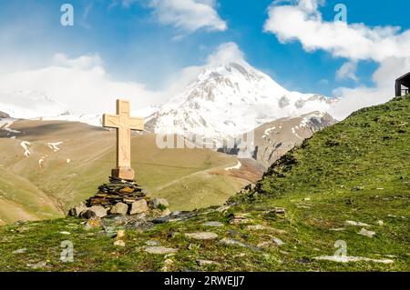 Photo of large cross standing on stones surrounded by greenery of high mountains in Kazbegi, Georgia Stock Photo