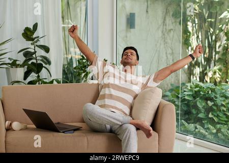 Young man sitting on couch and stretching after finishing work on laptop Stock Photo