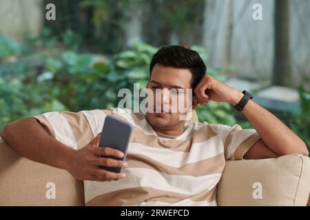 Portrait of young man sitting on couch and texting his friend or girlfriend Stock Photo