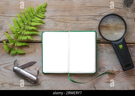 Blank notebook as mock up, fern frond, magnifying glass and a pocket knife on rustic wood, ecology concept for studying nature and environment, high a Stock Photo