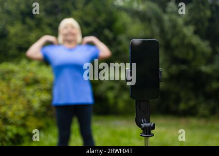 Fit mature 50s female in casual clothes exercising outdoors, practicing yoga on mat and talking at smartphone camera, making video content for Stock Photo
