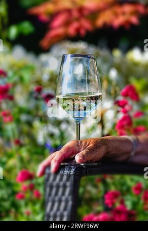 Close up of a glass of white wine resting on the arm of a garden chair with an soft focus colourful garden plants in the background on a summers day Stock Photo