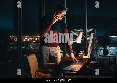 businessman standing at his desk in an office, talking on his mobile phone as he works late into the night. Professional man communicating with his co Stock Photo