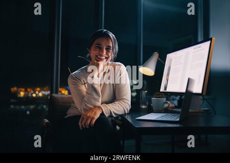 Happy business woman works late with her laptop and computer in a nighttime office. She plans and executes content marketing strategies after hours, s Stock Photo