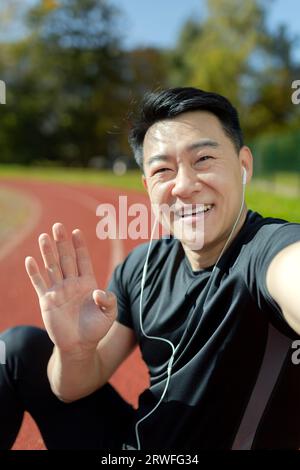 Close-up photo of a young Asian male athlete standing in a stadium wearing headphones, talking and greeting on a video call. Vertical photo. Stock Photo