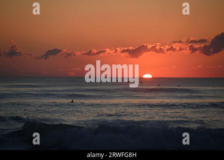 The sun rises on the horizon as distant surfers dot the water waiting to catch a wave in the Atlantic Ocean at Nags Head, North Carolina. Stock Photo