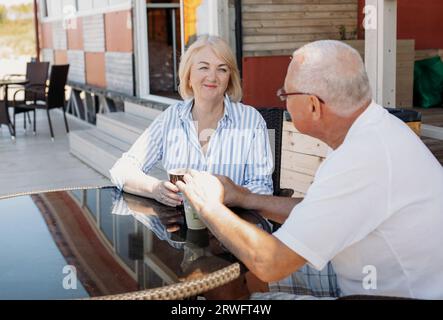 An elderly couple is sitting at a table in a cafe by the sea, looking at the sea, talking, drinking cocktails and coffee, holding hands. Stock Photo