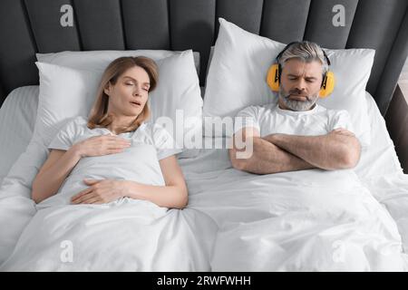 Irritated man with headphones lying near his snoring wife in bed at home Stock Photo