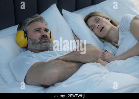 Irritated man with headphones lying near his snoring wife in bed at home Stock Photo